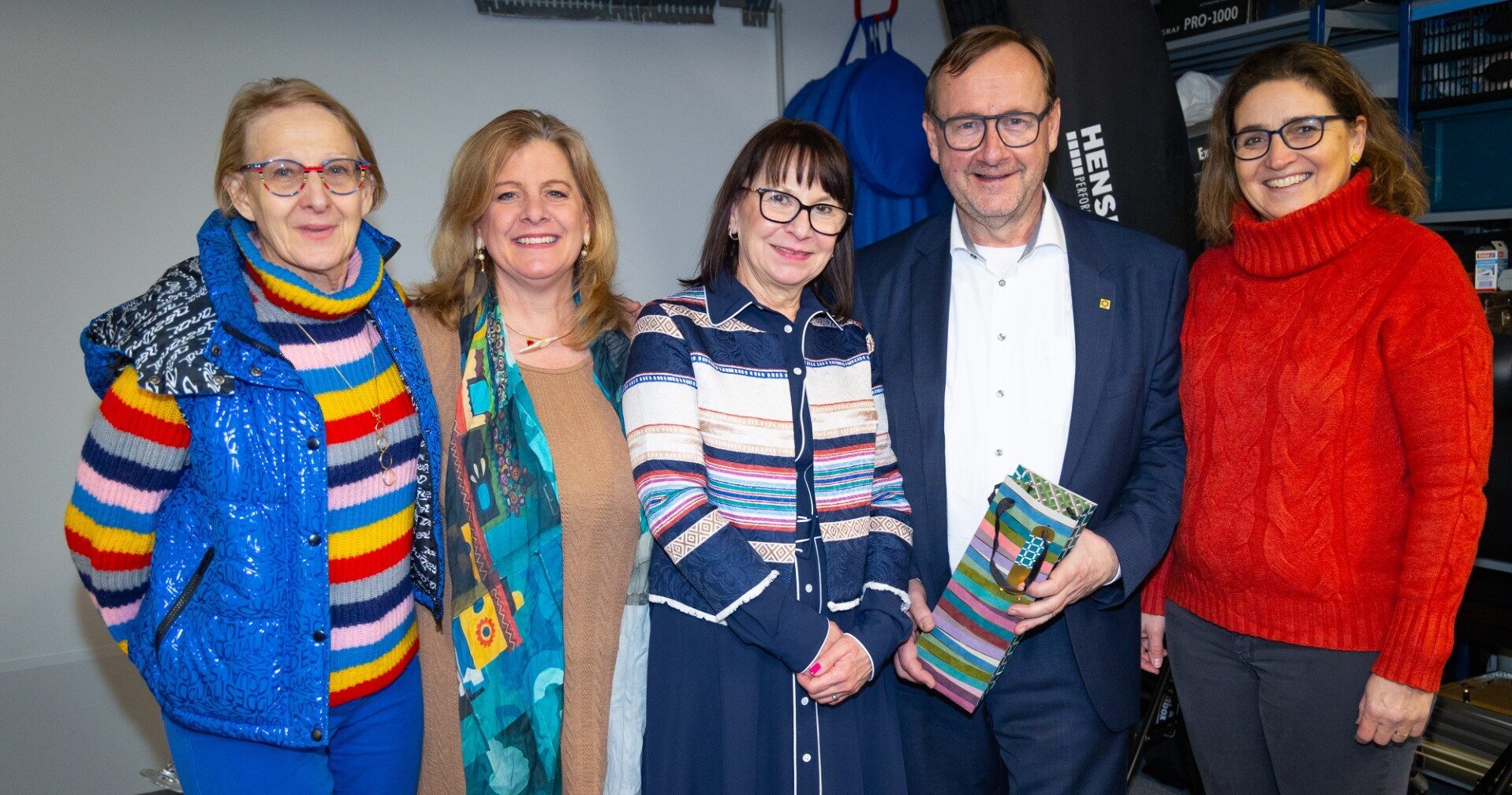 9ffb9a9f-0fad-4e0a-8327-b5196bcc3e19 Eine Gruppe von fünf lächelnden Personen – vier Frauen und ein Mann – steht nebeneinander in einem Innenraum. Der Mann in der Mitte trägt einen dunkelblauen Anzug und hält eine bunt gestreifte Geschenktüte. Die Personen tragen farbenfrohe Kleidung, darunter ein bunter Rollkragenpullover und ein roter Strickpullover.